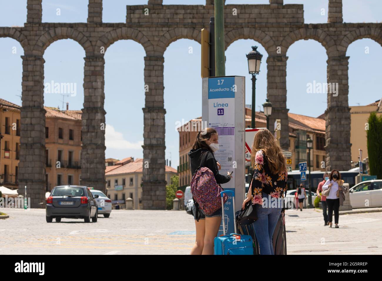 Segovia, Spain - June 2, 2021: Two young girls wait at a bus stop with their suitcases, near the Aqueduct of Segovia, to go to the train station Stock Photo