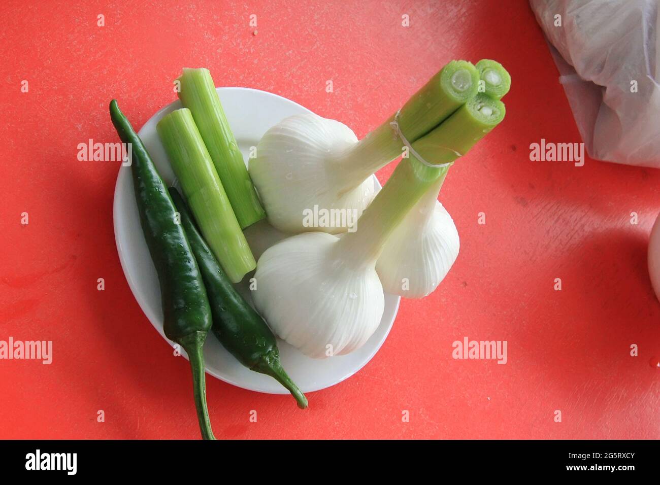 Three heads of young garlic and two green chili peppers in a white plate on a red table Stock Photo