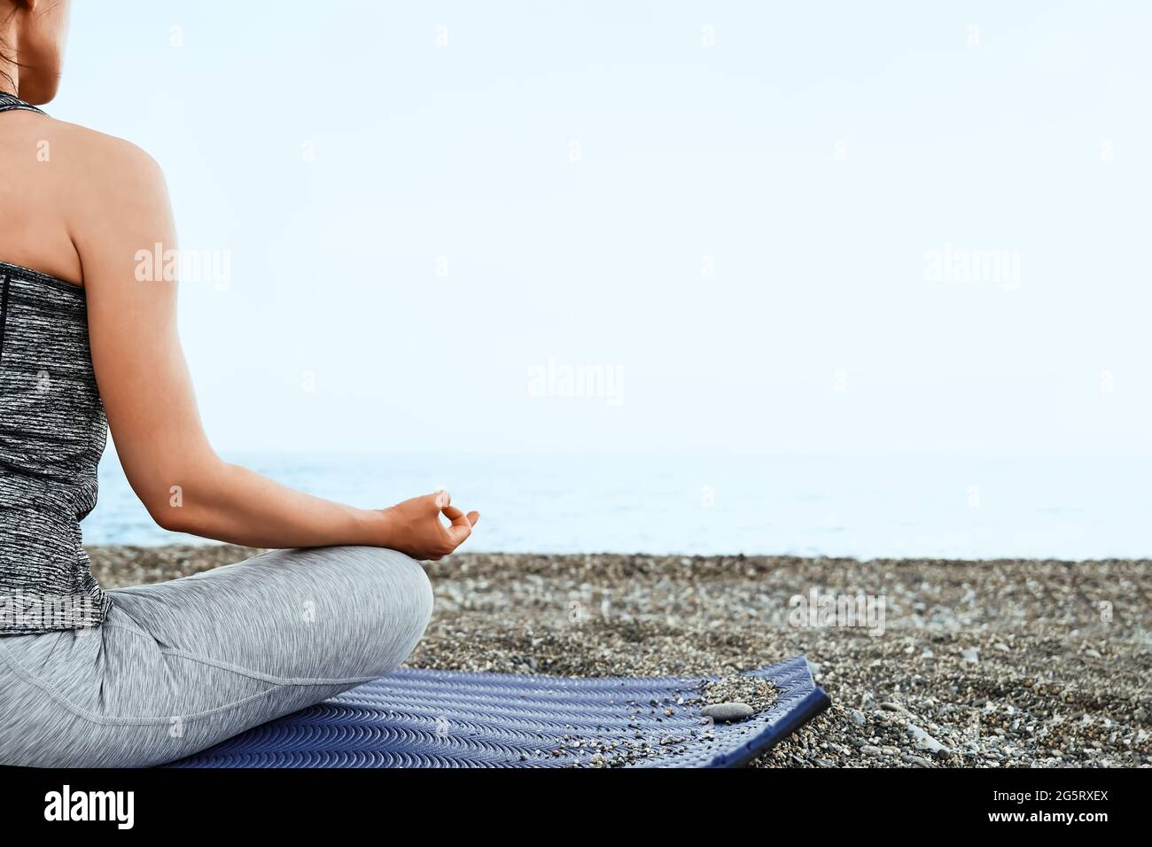 Woman sitting in lotus poses practices yoga on the beach in the morning, she's meditating, looking on the blue sea. Wellbeing concept. Stock Photo
