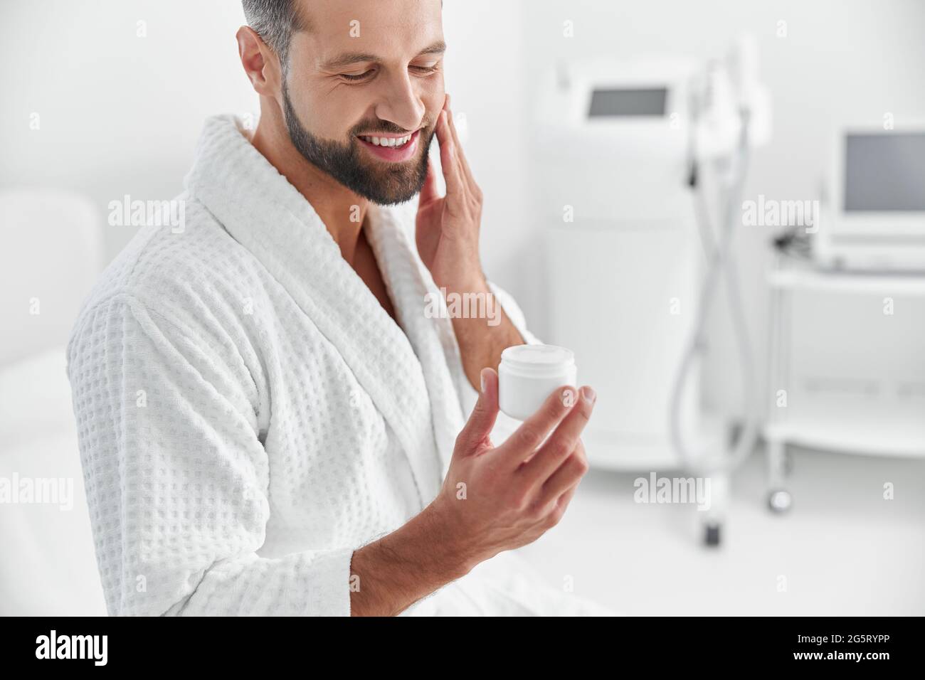 Smiling mature man applies cream onto cheek in modern cosmetologycal salon Stock Photo
