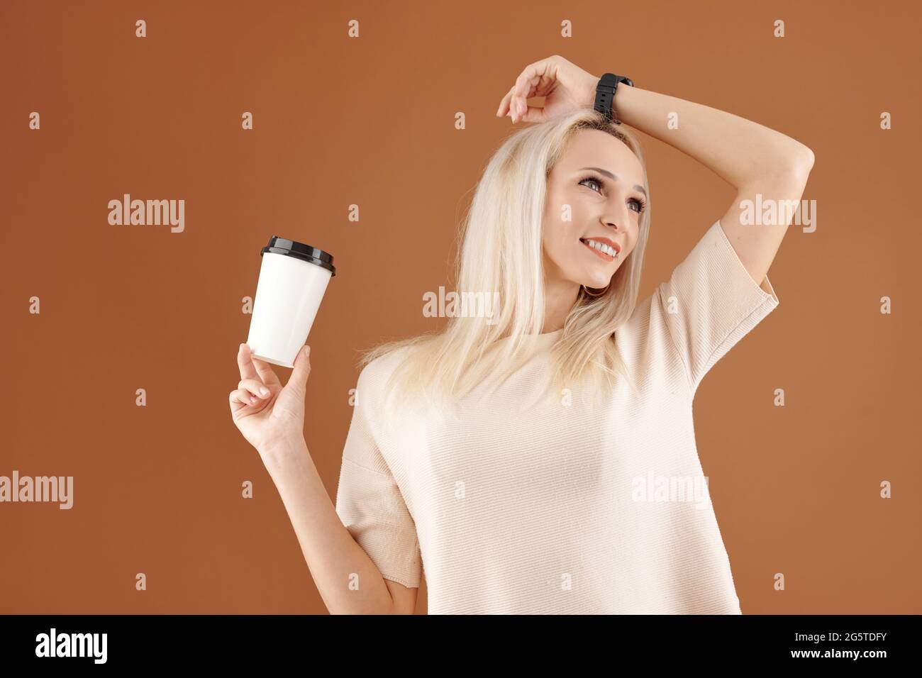 Happy carefree young blond-haired woman in wristwatch holding takeout coffee cup and holding hand on head while looking away Stock Photo