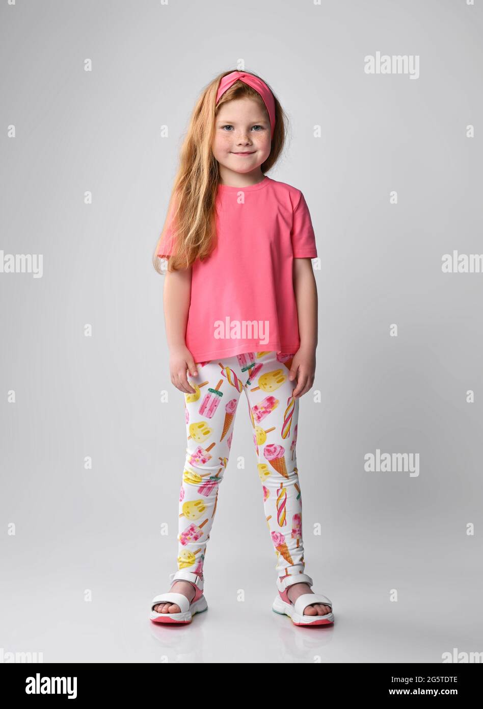 Cute smiling red-haired kid girl stands in summer girlish wear pink t-shirt, colorful pants with print and sandals Stock Photo