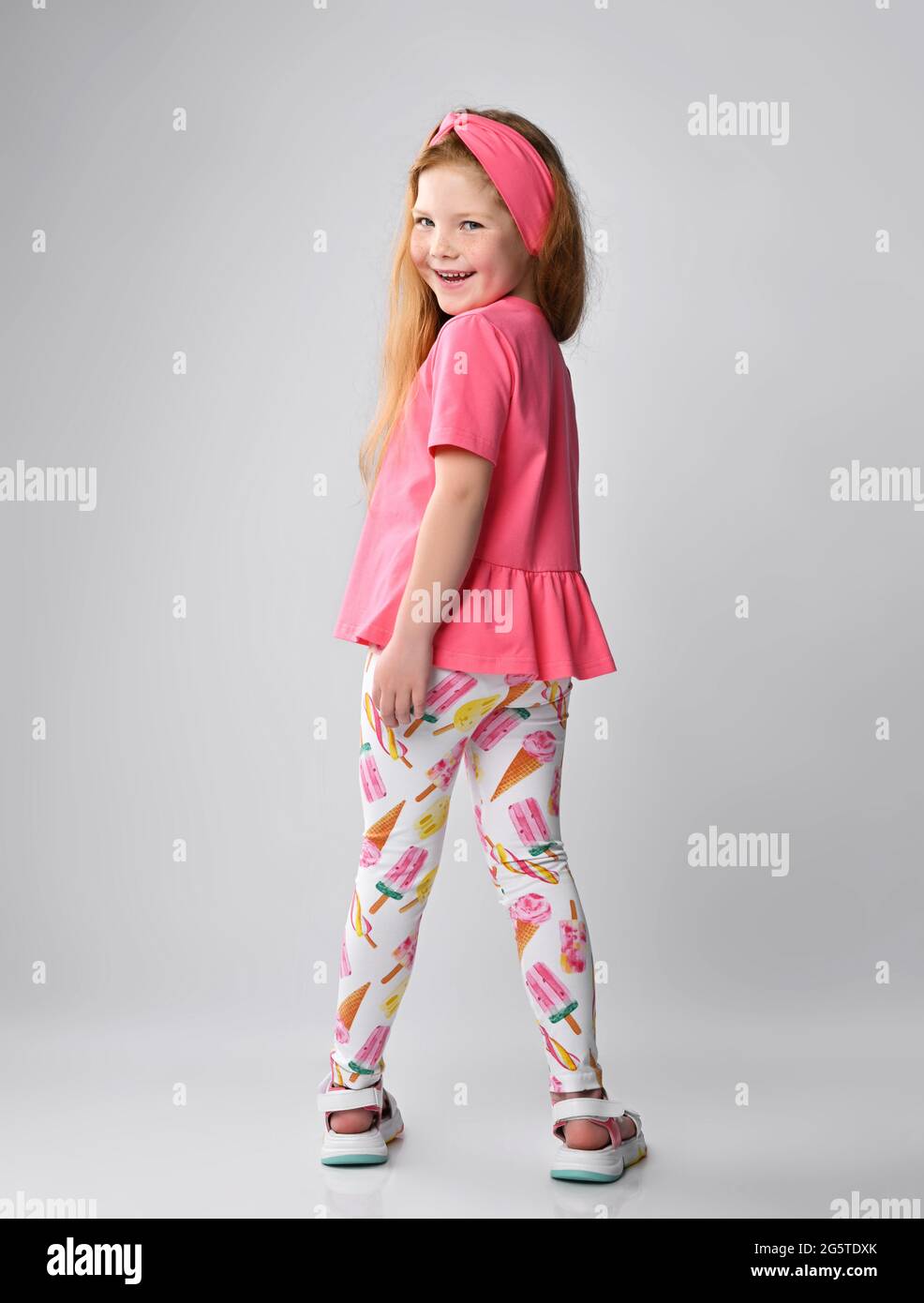 Smiling red-haired kid girl in pink t-shirt, colorful pants and sandals stands with her back to camera and turned Stock Photo