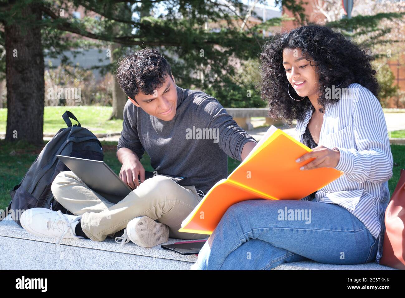 Two latin students studying from their lecture notes sitting on a wall outdoors. University life at campus. Stock Photo