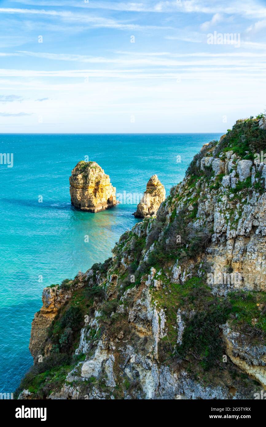 Rocky cliffs with fresh green vegetation and sunlit sea stacks in the turquoise ocean at the coast of Lagos, Portugal - vertical orientation Stock Photo
