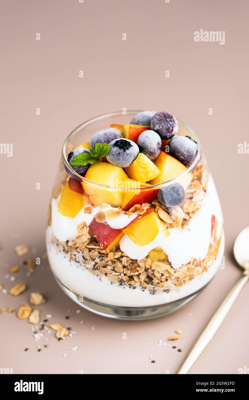 Greek yogurt, granola and fruits in jar. Healthy vegetarian breakfast parfait with peach, blueberries, oat honey granola and yogurt, beige background Stock Photo