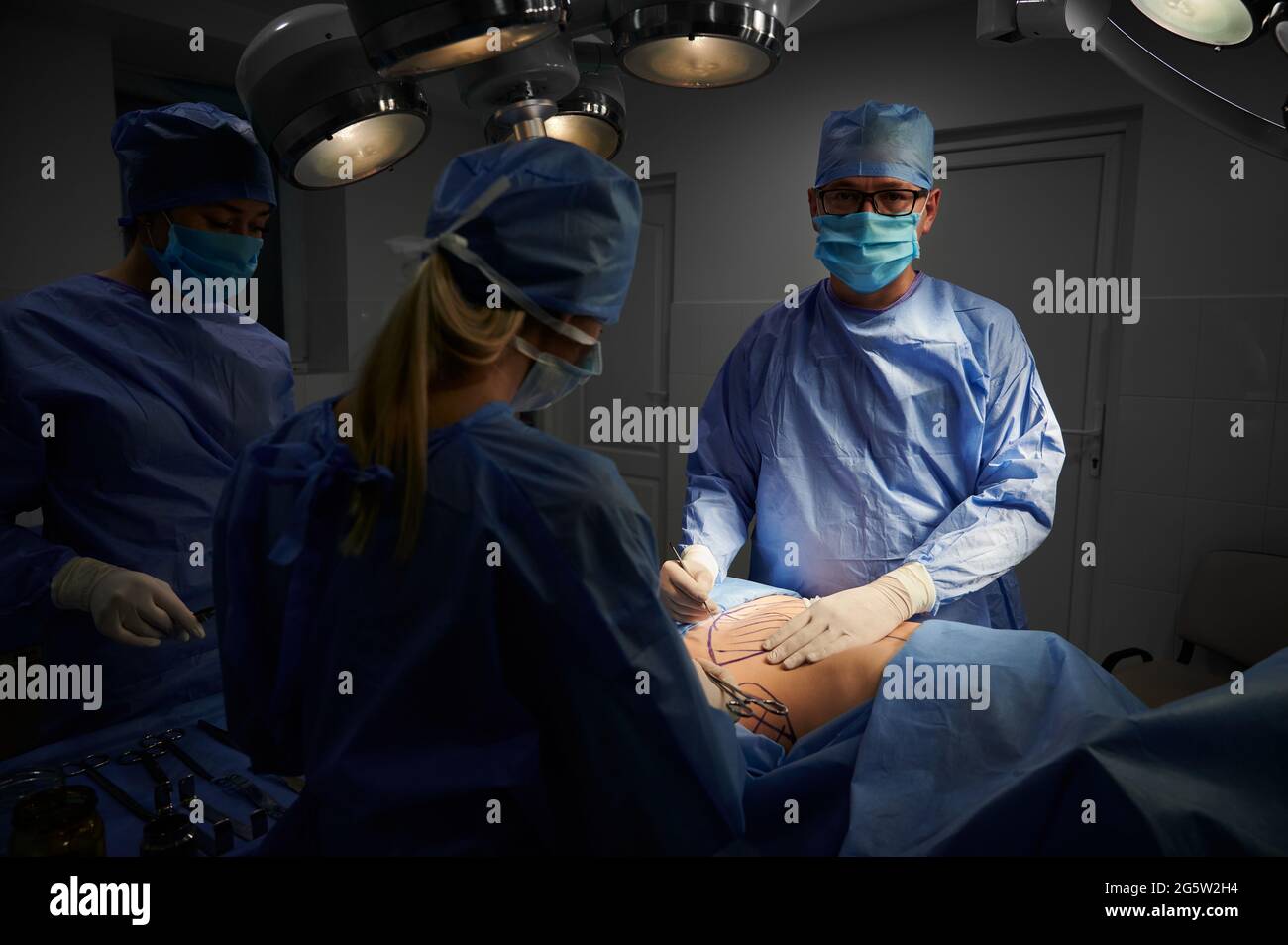 Medical team doing aesthetic plastic surgery in operating room with dim light. Surgeon performing cosmetic surgery, looking to the camera. Concept of medicine and plastic surgery Stock Photo