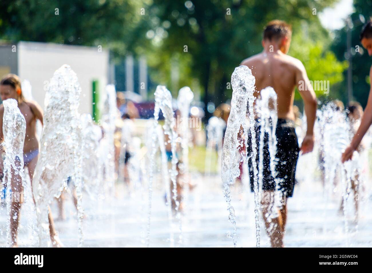 Selective focus on jets of water in a city fountain on a hot summer day. In the blurred background children and adults are batning and having fun. Stock Photo
