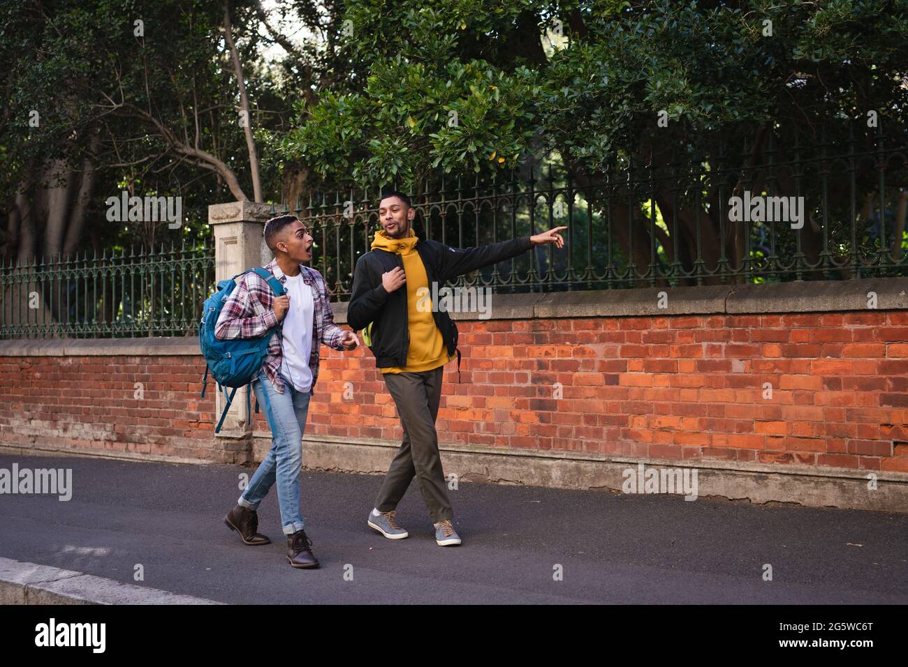 Two happy mixed race male friends carrying backpacks walking in city street talking, one pointing Stock Photo