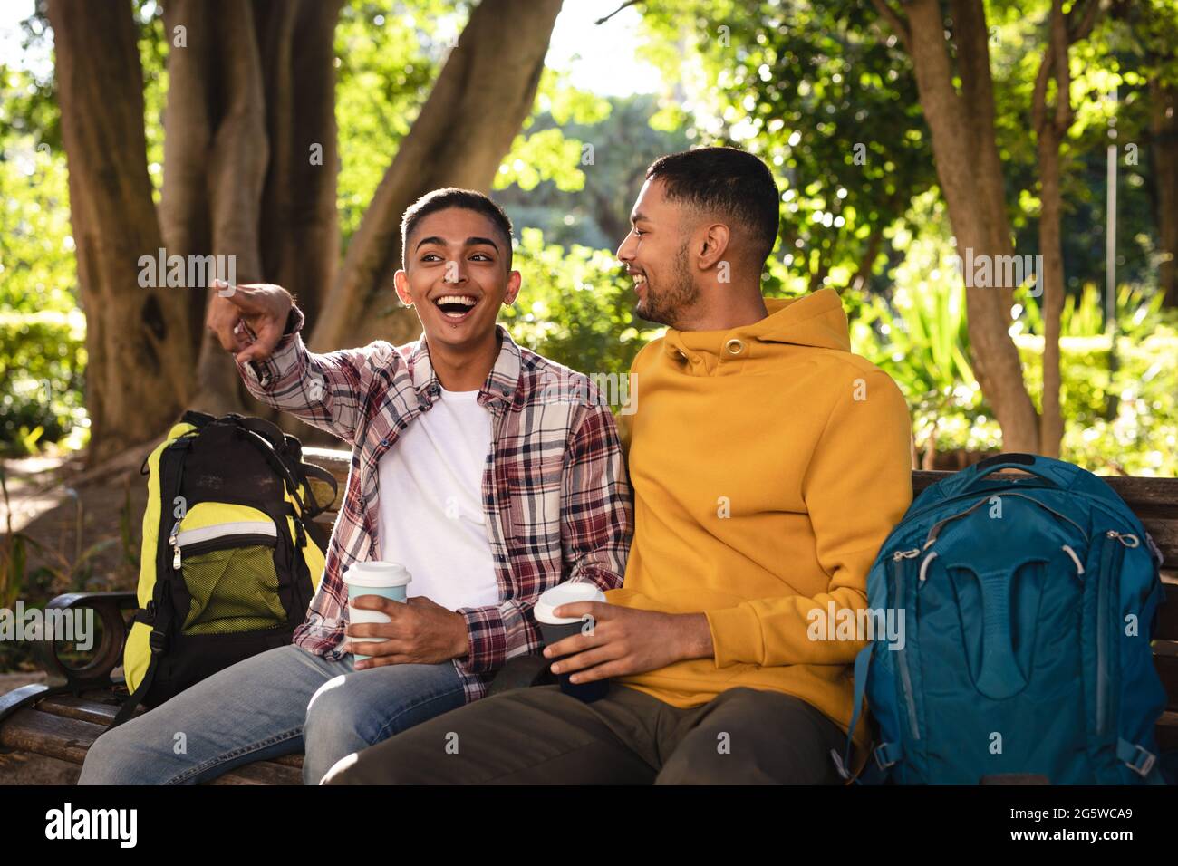 Two happy mixed race male friends sitting on park bench with backpacks, talking Stock Photo