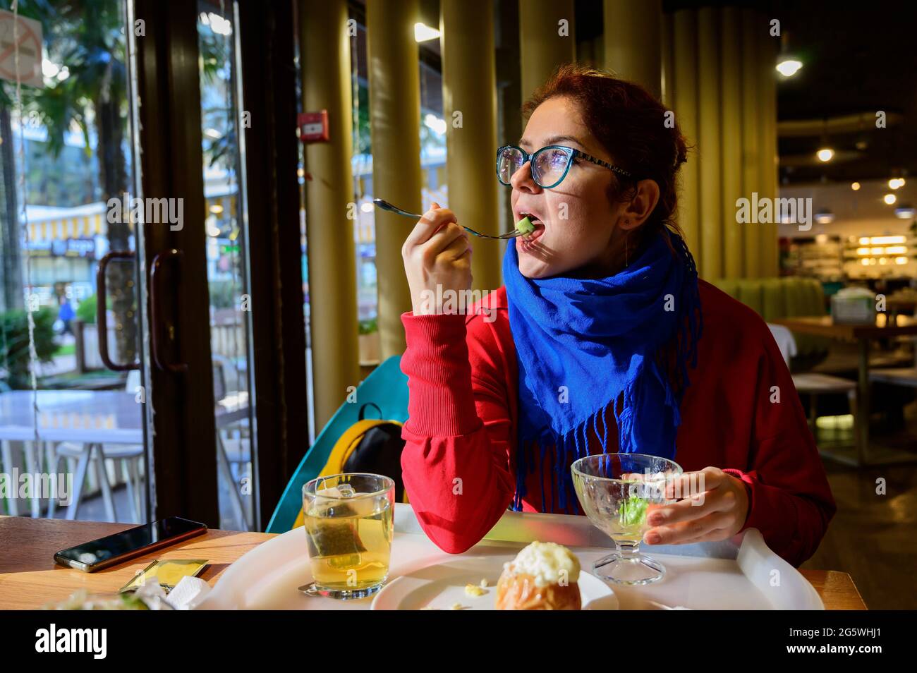 Red-haired caucasian woman eating salad in a cafe. Pensive romantic girl in  glasses and a blue scarf is having lunch Stock Photo - Alamy