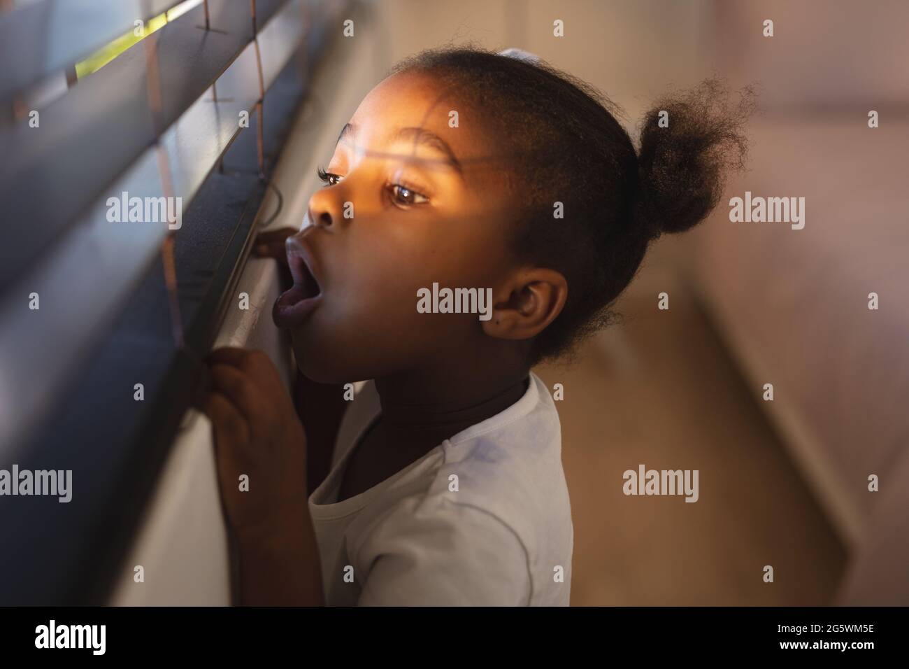 Surprised african american girl standing and peering through window blinds on a sunny day Stock Photo