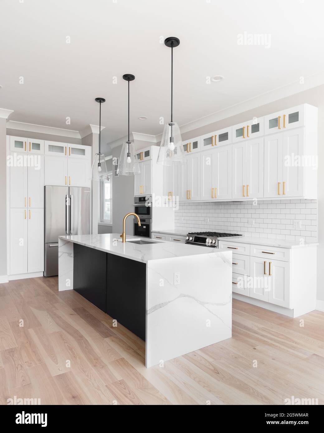 A luxury white kitchen with black pendant lights hanging above a waterfall granite island, stainless steel Bosch appliances, and gold hardware. Stock Photo