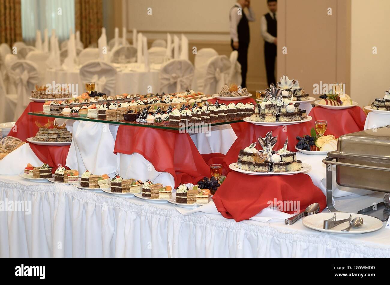 Catering table with a red and white tablecloth with a lot of sweet snacks, small cakes with cream and chocolate cream in a large banquet hall in the b Stock Photo