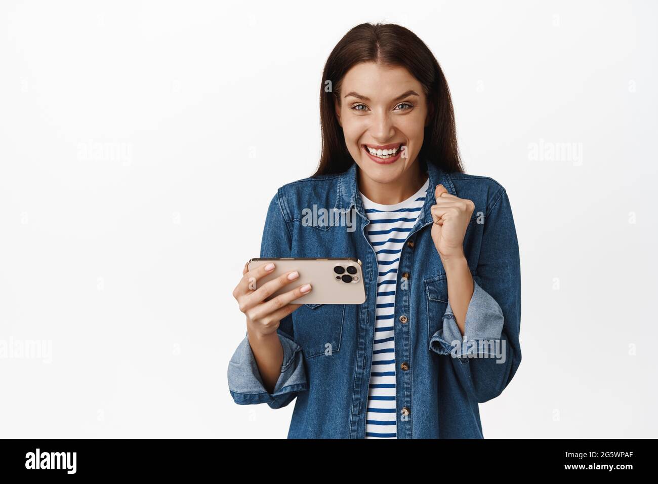 Happy brunette woman winning money on phone, playing mobile video game and rejoicing, celebrating goal achievement in app, standing against white Stock Photo
