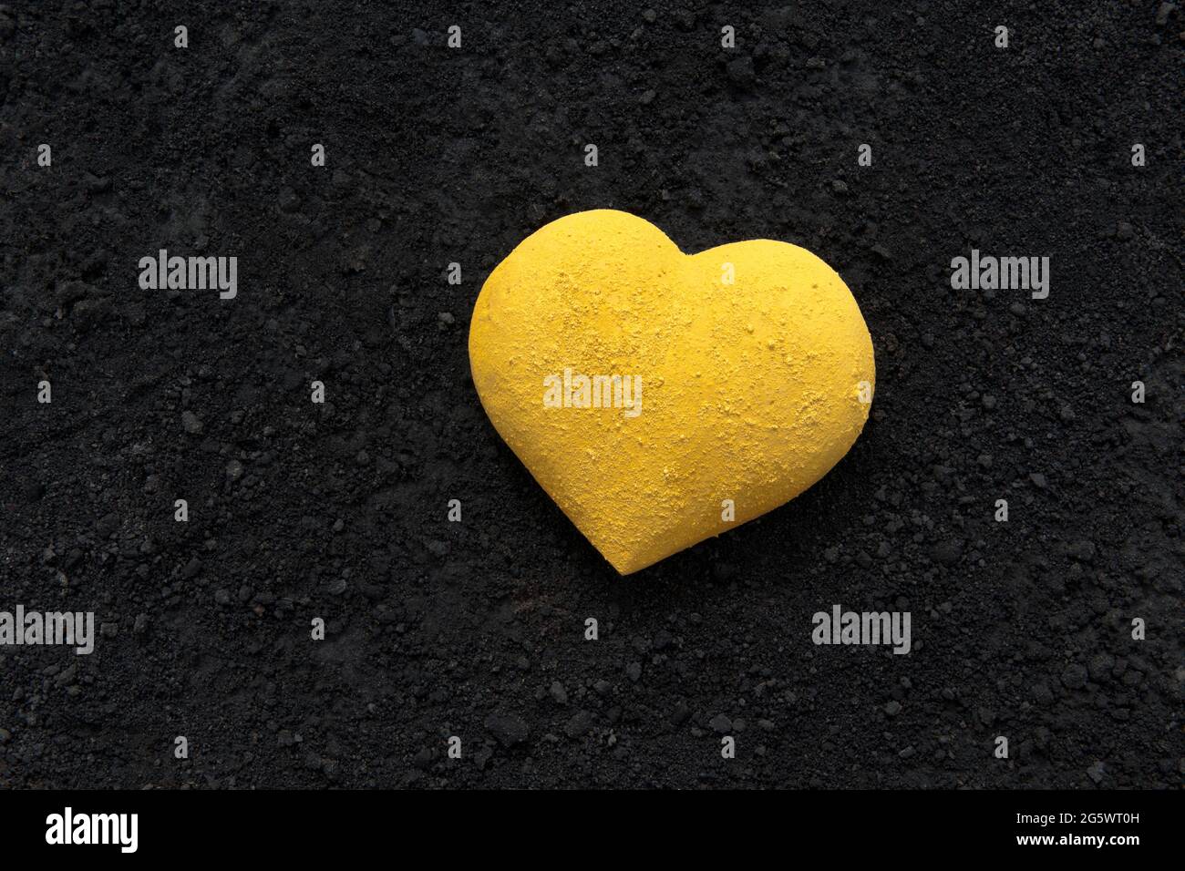 3 dimensional heart shape painted bright yellow with powder paint, placed on a background of black cinders Stock Photo