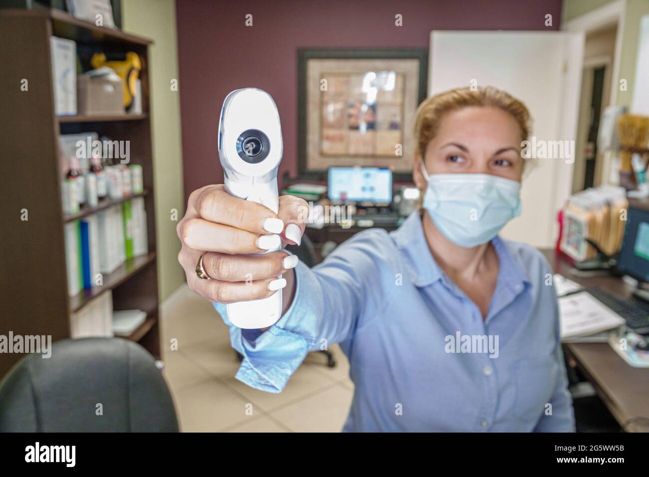 Miami Beach Florida dental office temperature check receptionist Hispanic female Covid-19 pandemic wearing face mask points non-contact IR thermometer Stock Photo