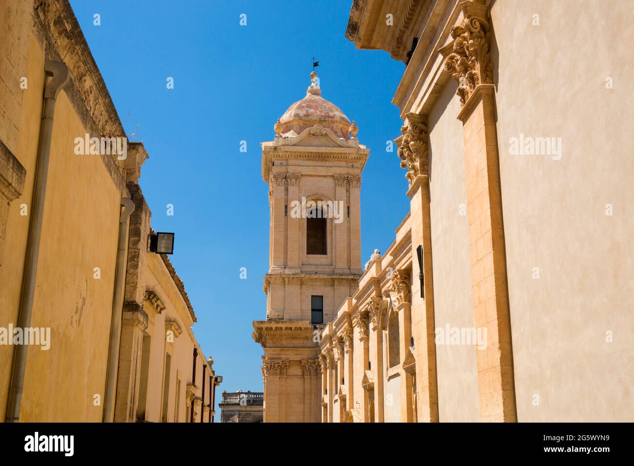 Noto, Syracuse, Sicily, Italy. View along sunlit narrow street to clock-tower of the baroque Cathedral of San Nicolò. Stock Photo