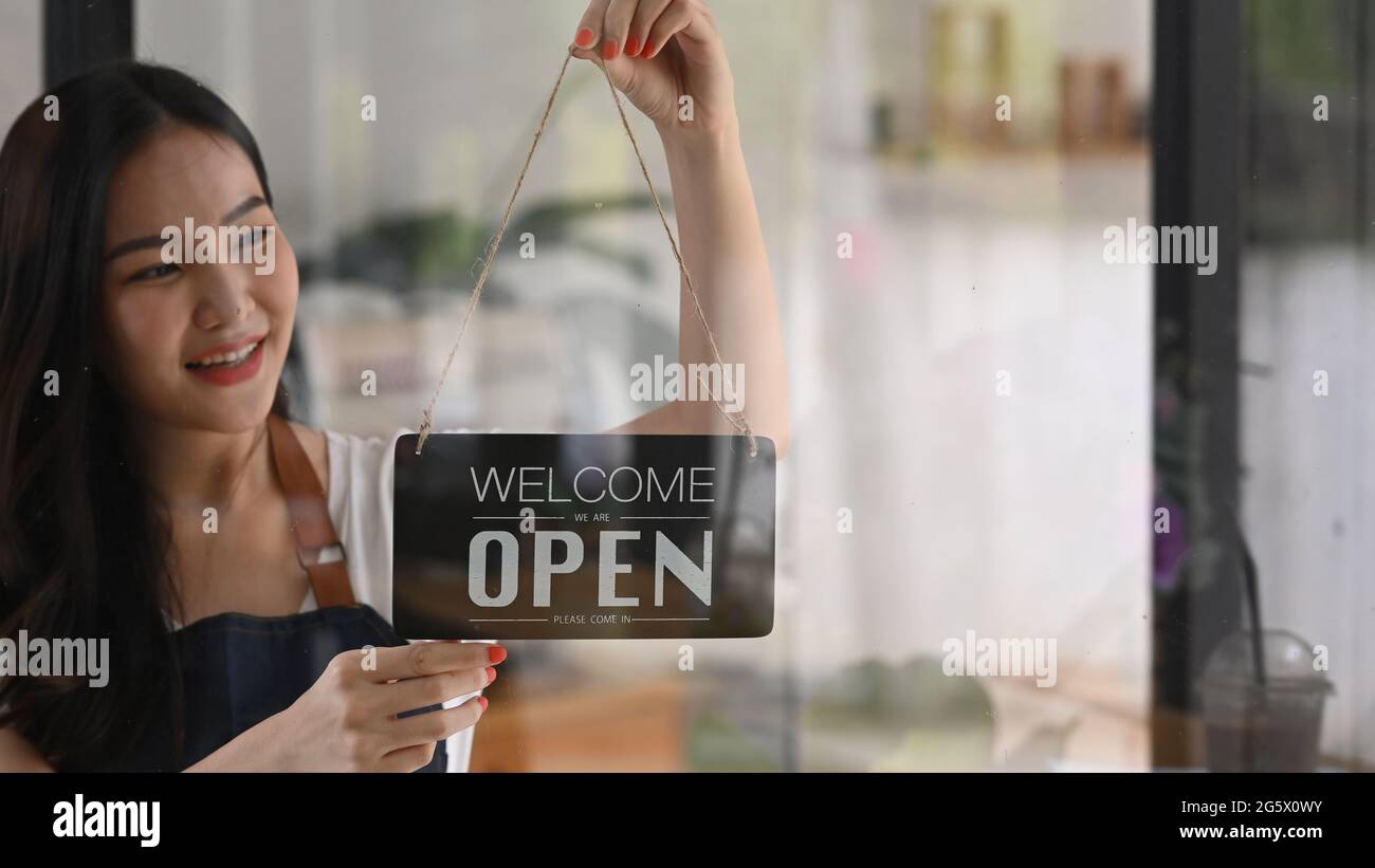 Smiling woman owner turning open sign board on glass door in modern coffee shop. Stock Photo