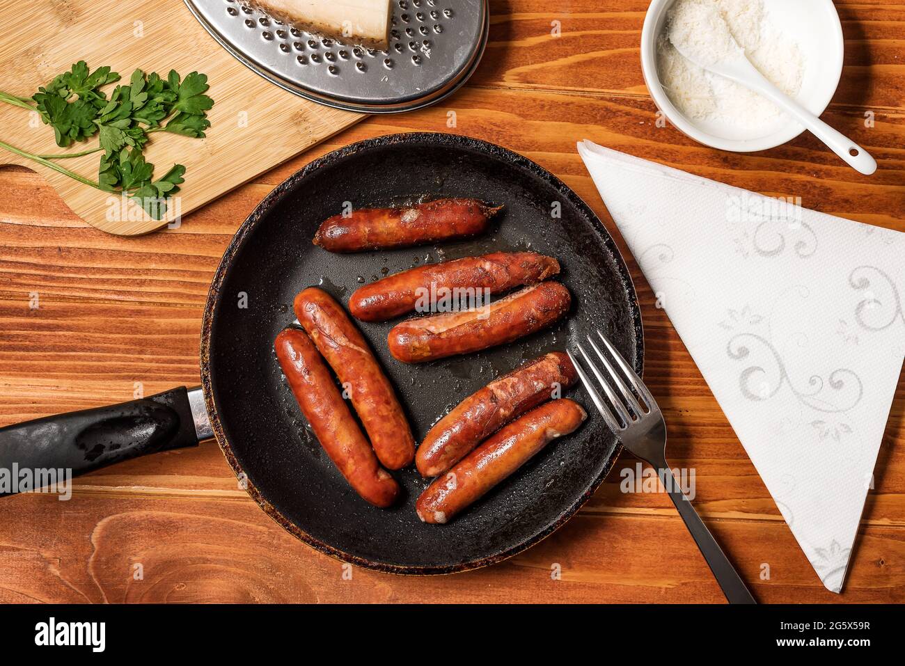 Tasty fried sausages in frying pan with Parmigiano and parsley, over wooden background. Top view Stock Photo