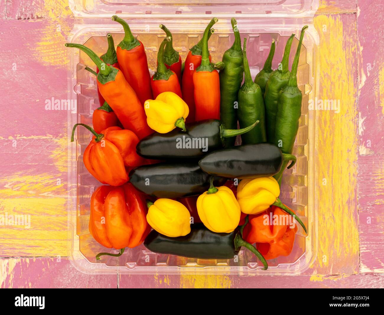 Plan view of assorted chilli peppers in a clear plastic carton, on a pink and yellow wooden rustic background. Stock Photo