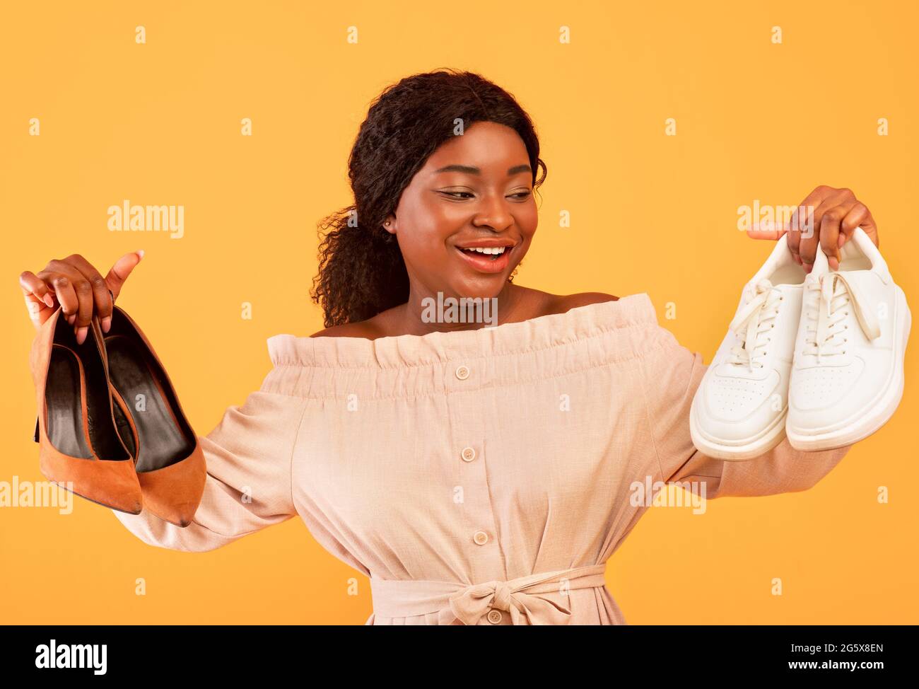 Millennial black female choosing between elegant high-heeled shoes and comfy sneakers on orange studio background Stock Photo