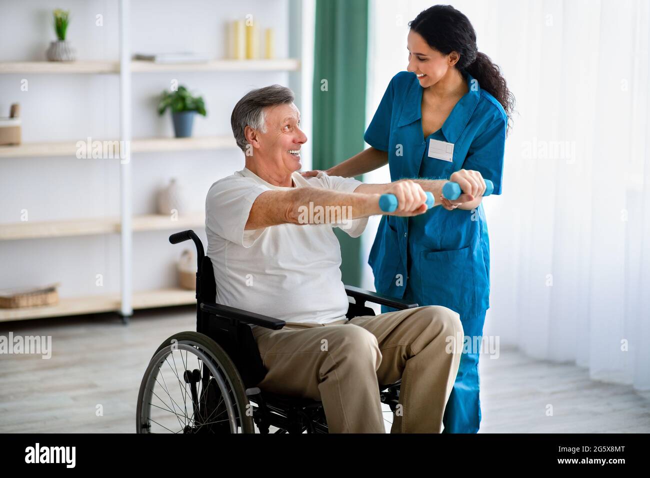 Female physiotherapist helping elderly man in wheelchair do exercises with dumbbells at health centre Stock Photo