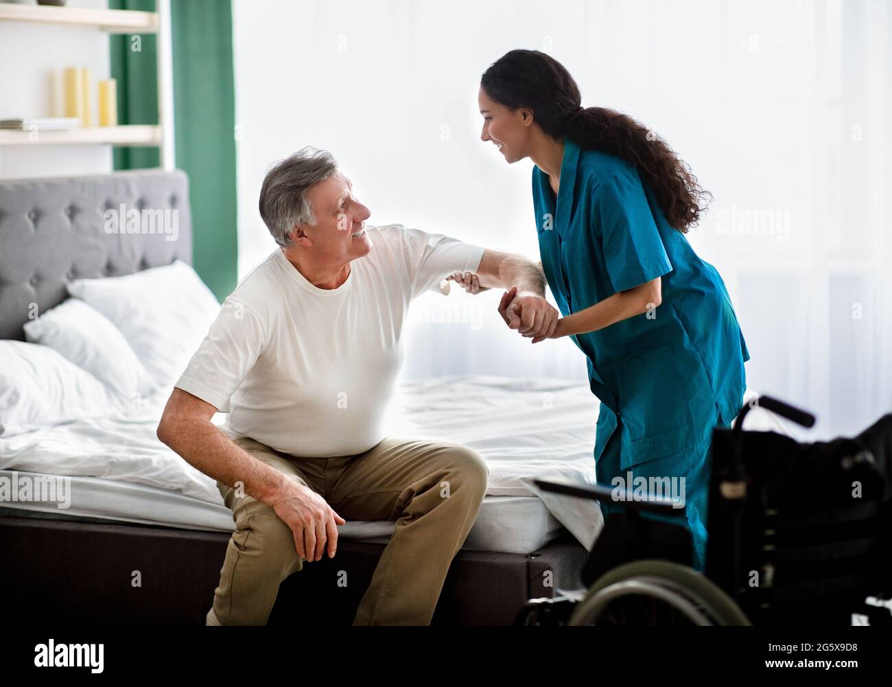 Caregiver helping elderly male to get into wheelchair at home. Rehabilitation and medical care for disabled seniors Stock Photo