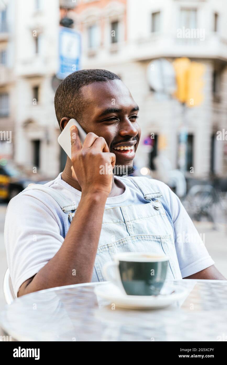 vertical portrait of an afro american young man. He is talking by phone and smiling at a bar terrace Stock Photo