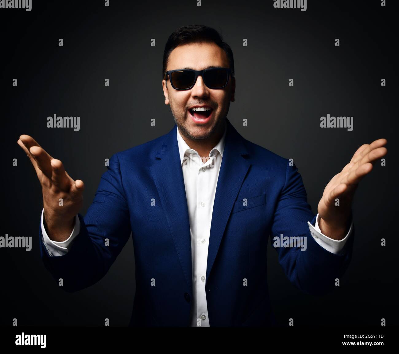 Happy laughing brunet man in blue jacket, white shirt and sunglasses stands holding hands with open palms up at camera Stock Photo