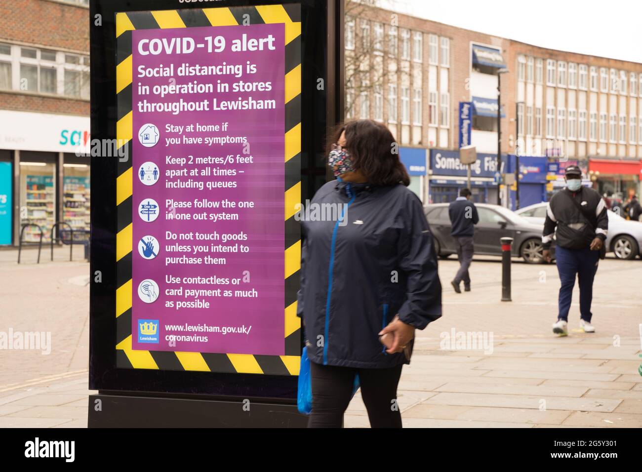 COVID-10 alert message on giant LED screen in high street Lewisham , London, England, Europe, woman wears face mask with her shopping in hand Stock Photo