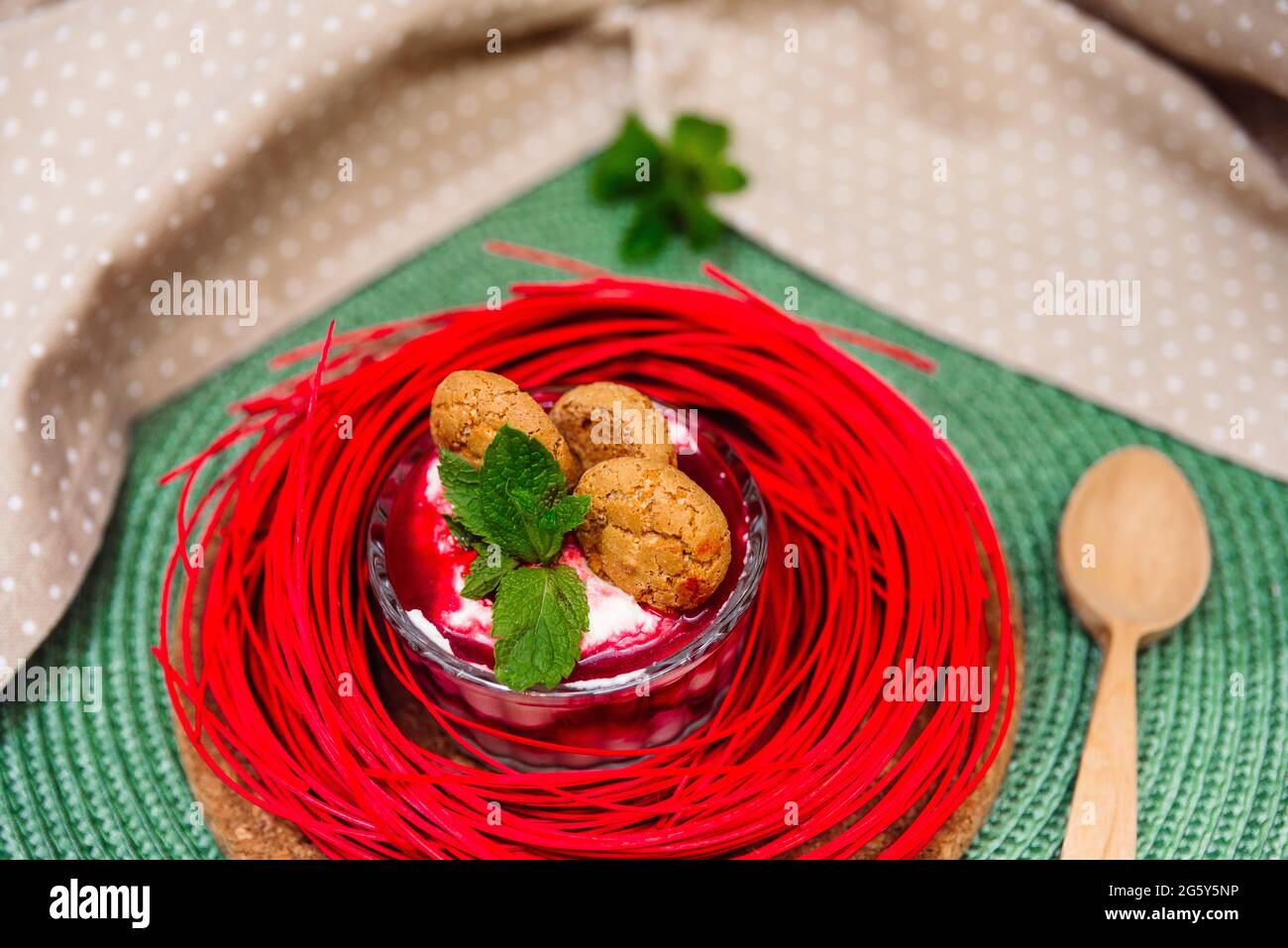 Dessert parfait with strawberry jam in a glass jar decorated with cracked cookies, mint leaves on a wicker green napkin. One small home food portion. Stock Photo