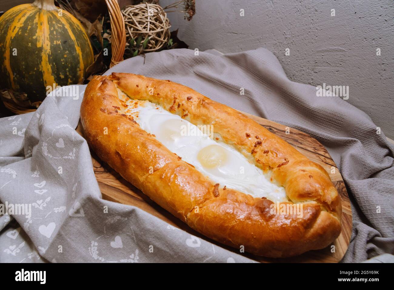 Baked Adjarian-style hot khachapuri with eggs and melted cheese right from the oven are resting on wooden board with a grey towel to the right Stock Photo