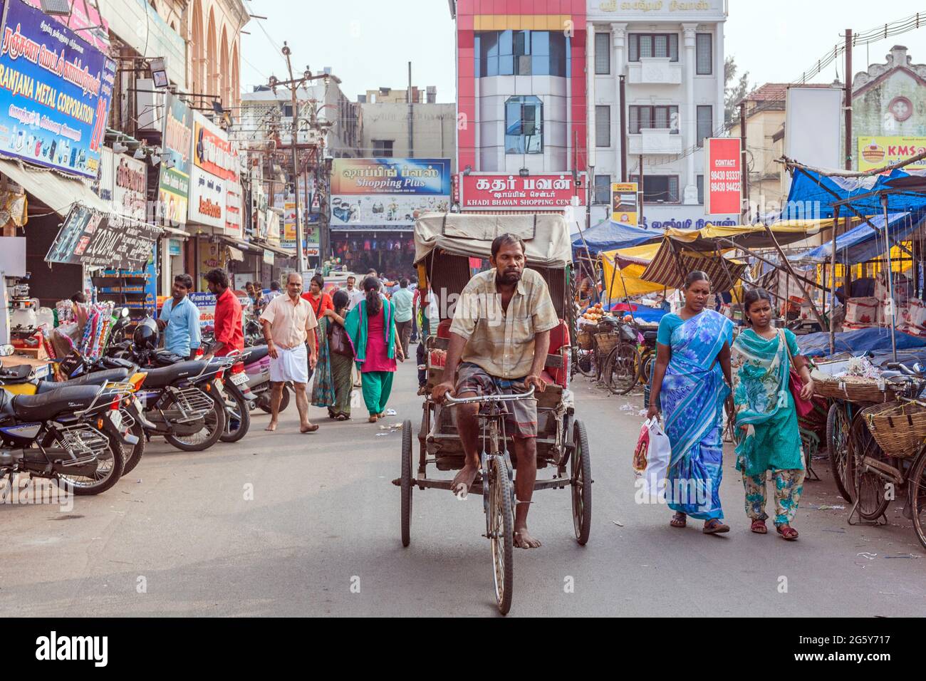 Cycle Rickshaw cycles through busy city centre with pedestrians, market and shopfronts, Trichy, Tamil Nadu, India Stock Photo