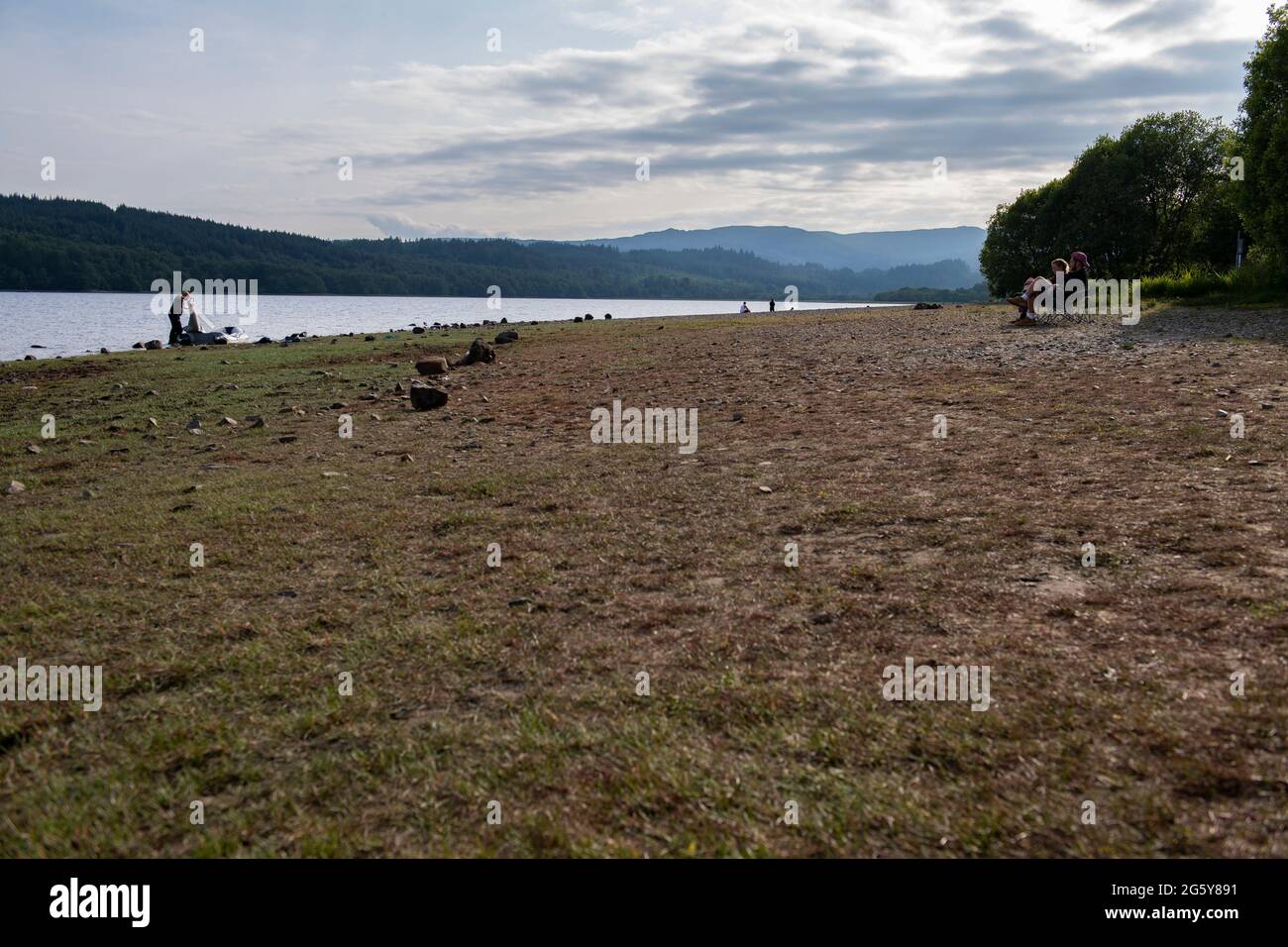 Loch Venachar, Loch. Lomonnd and Trossachs National Park, Scotland, UK. 30th June, 2021. PICTURED: A wide angle view of the pebble and stone beach on Loch Venachar which is normally under a few feet of water, is seen exposed due to the dwindling water supply being used up. There should not be any beach on this part of the loch, however people can be seen using this make shift beach to take in the evening sun shine. Credit: Colin Fisher/Alamy Live News Stock Photo