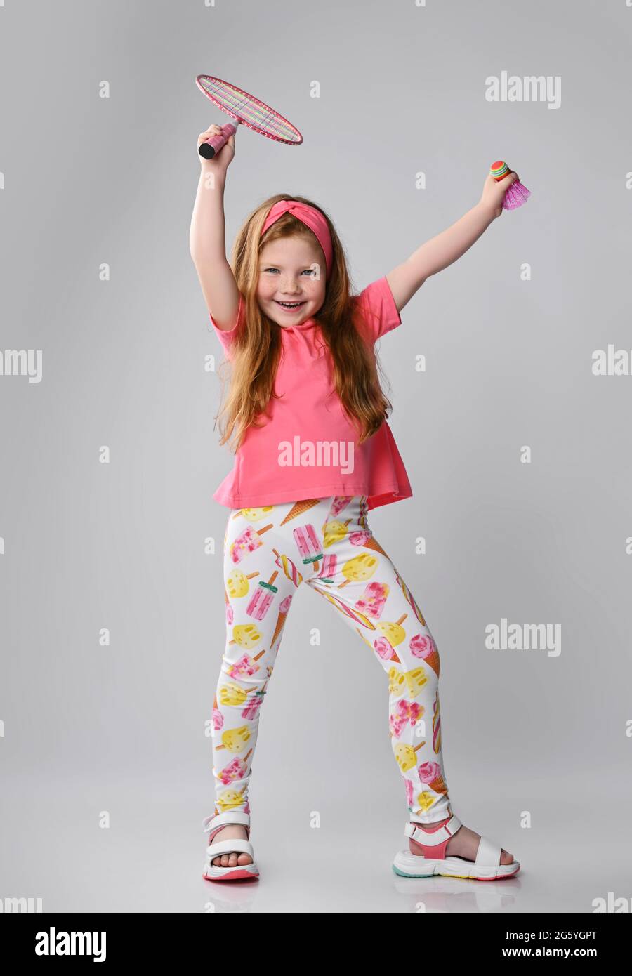 Happy screaming red-haired kid girl in colorful summer clothes stands waving her badminton racquet and shuttlecock Stock Photo