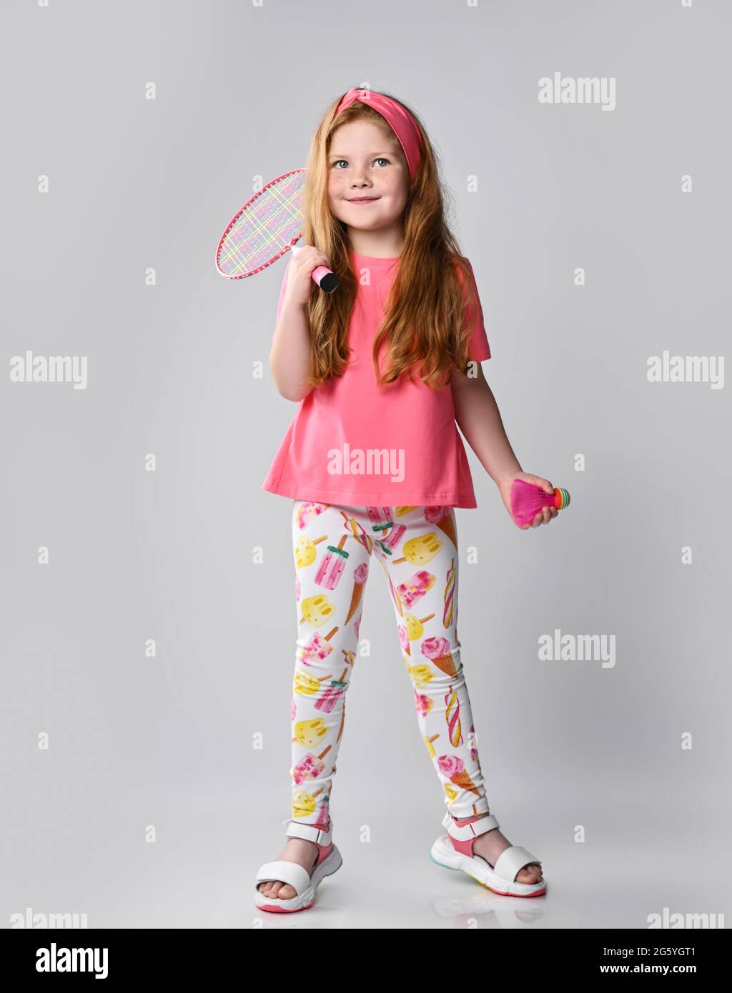 Smiling redhead kid girl in colorful summer clothes holds a badminton racquet on her shoulder and shuttlecock in hand Stock Photo