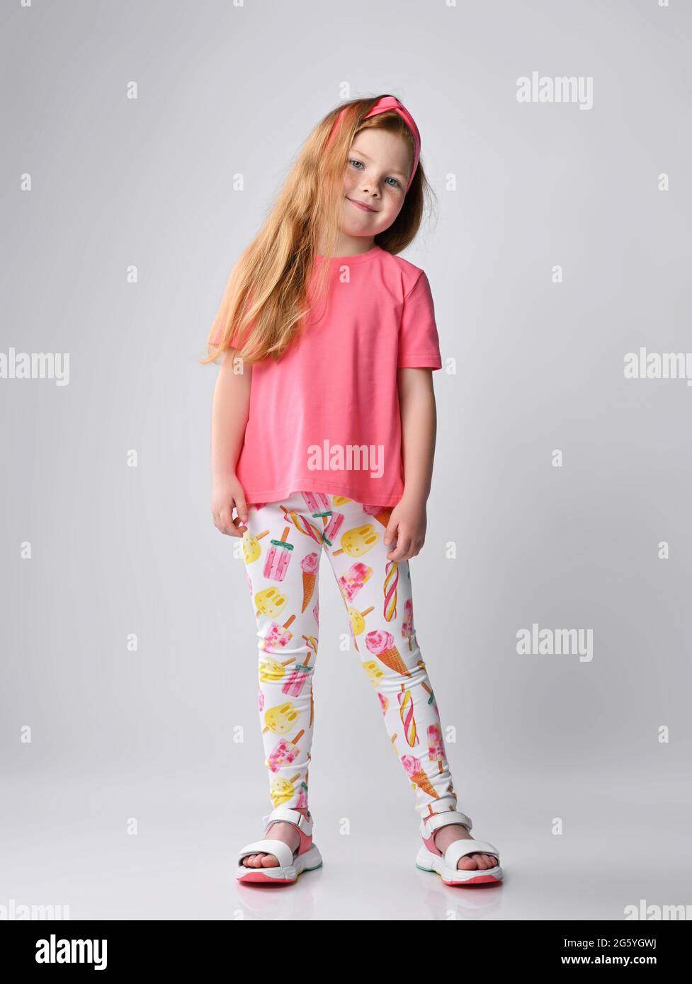 Red-haired kid girl in summer girlish clothing, wear pink t-shirt, colorful pants with ice-cream print and sandals Stock Photo