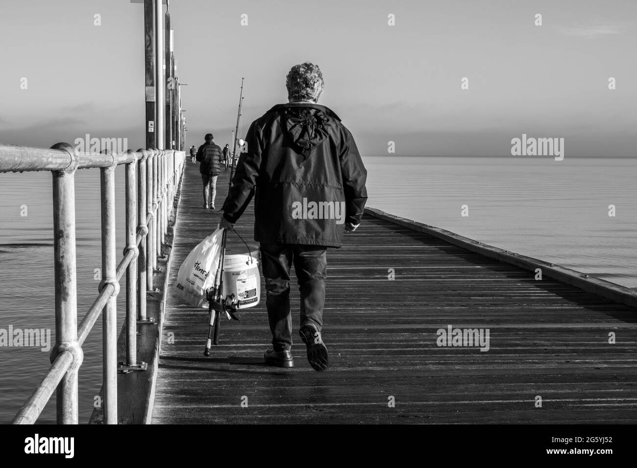 Fisherman walking along a wooden pier on a calm sea with fishing rods and bucket in Frankston Victoria Australia Black and white Stock Photo