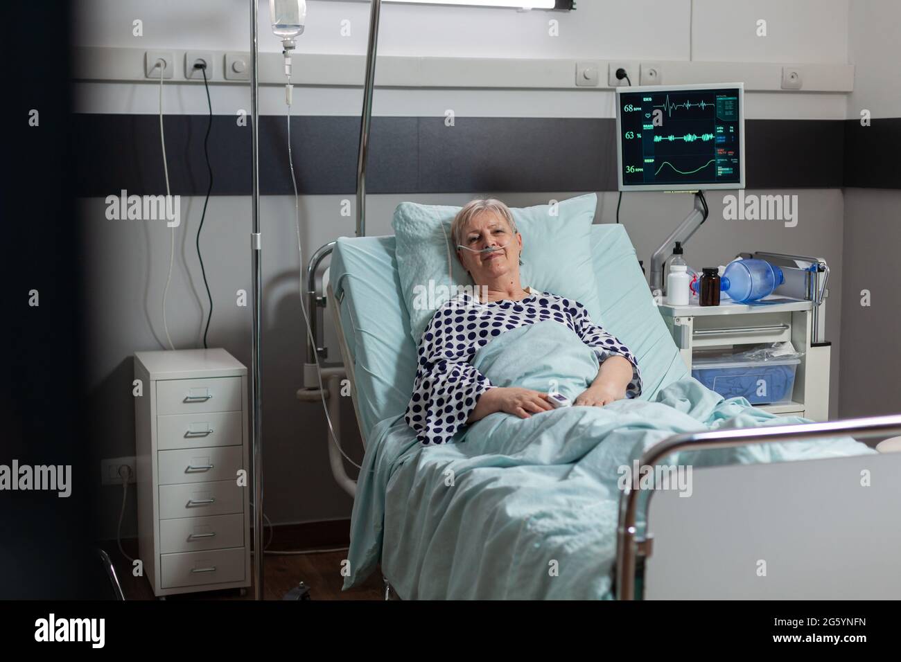 Portrait of senior woman smiling looking at camera laying in hospital bed betting treatment through iv drip bag. Breathing with help from oxygen mask during illness recovery. Stock Photo