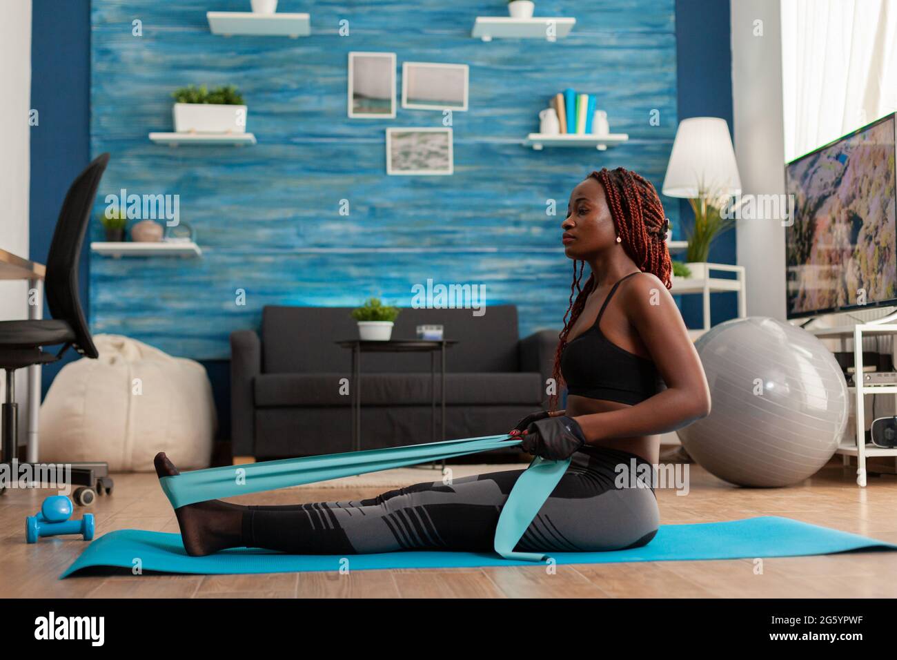 Black woman doing pilates workout using elastic strap sitting on yoga mat,  pulling training arms and shoulders in home living room. Athletic fit  exercising body using resistance band Stock Photo - Alamy