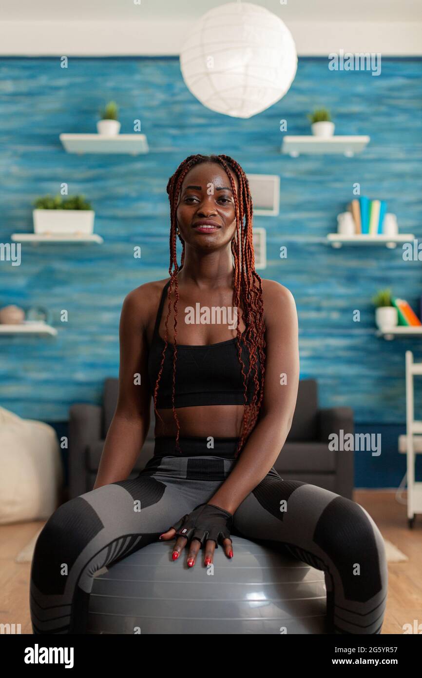 Happy joyful smiling african woman relaxing on swiss ball, after intense hard sport workout on yoga mat in home living room. Cheerful strong athletic fit african using stability ball. Stock Photo