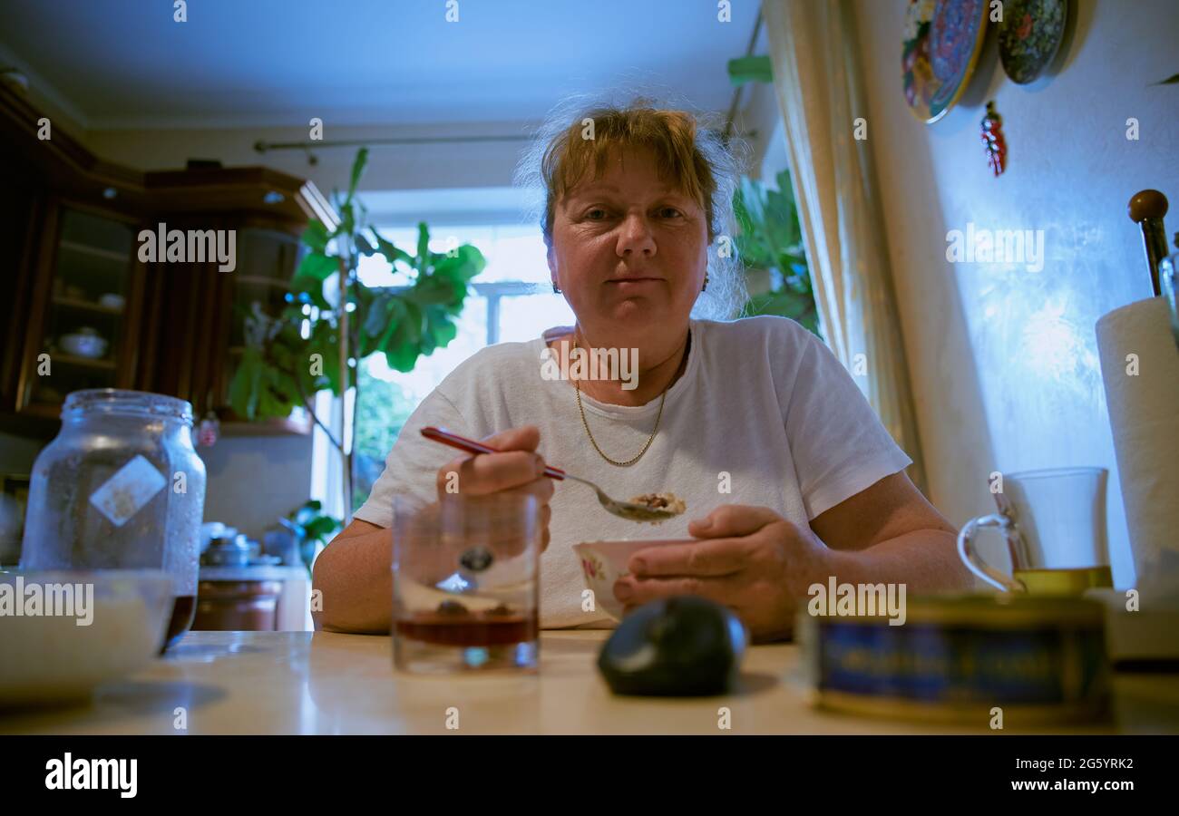Blonde adult lonely woman sitting in the kitchen at the table while eating with plants on background, Close- up photo, natural light, color. Stock Photo