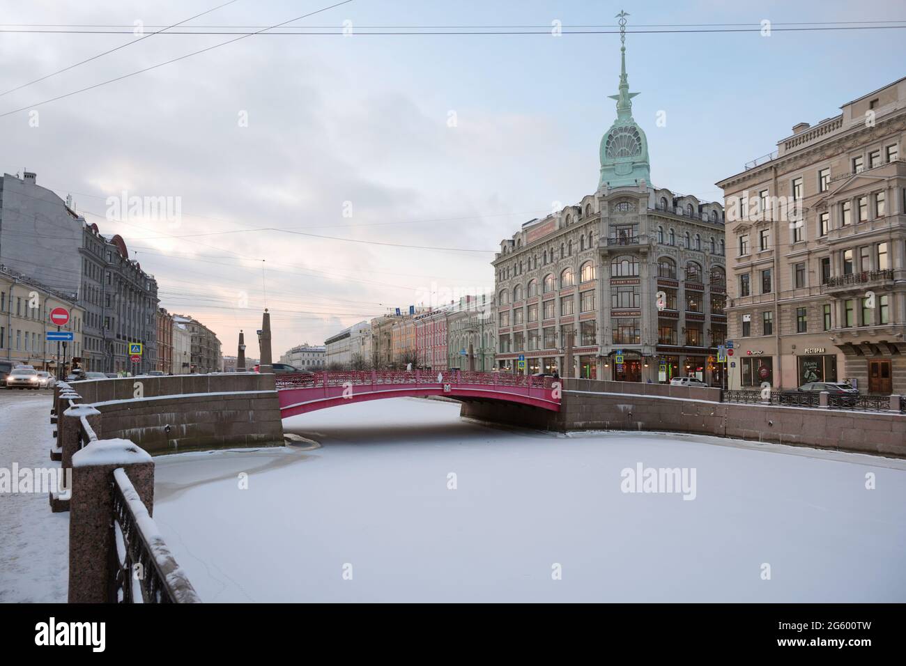 St. Petersburg, Russia, 16th January, 2021: Red Bridge across frozen river Moika with the art nouveau building of shopping centre At The Red Bridge, Red Bridge was built in 1808-1813 by design of William Heste, and the building of shopping centre was erected in 1906-1907 for the clothing store of Stefan Esders and Charles Scheefhals by design of architects Vladimir Lipsly and Konstantin de Rochefort Stock Photo