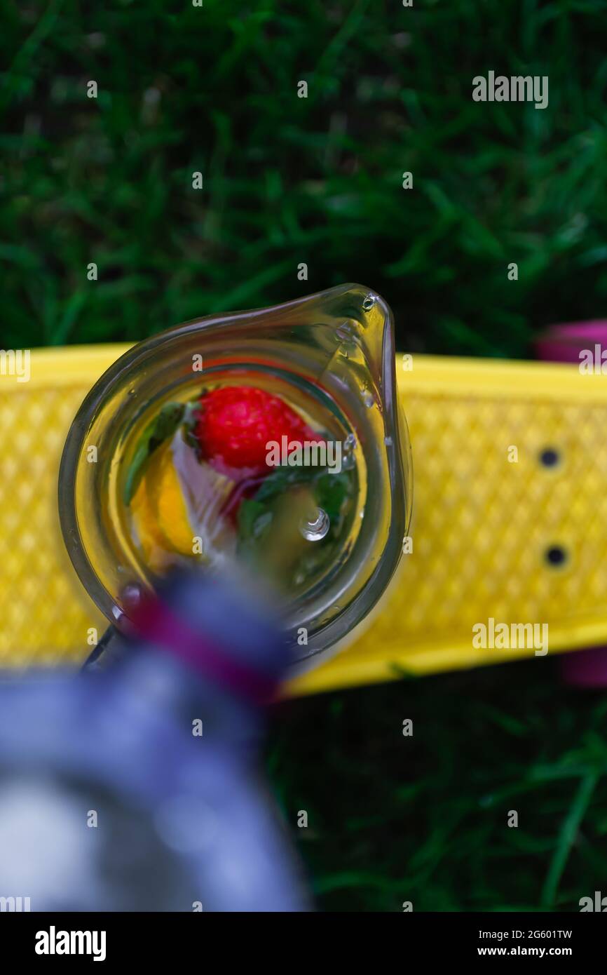 Defocus drop of water pouring from plastic bottle in glass jug of lemonade with strawberry, slice lemon and leaves of mint on yellow penny board, skat Stock Photo