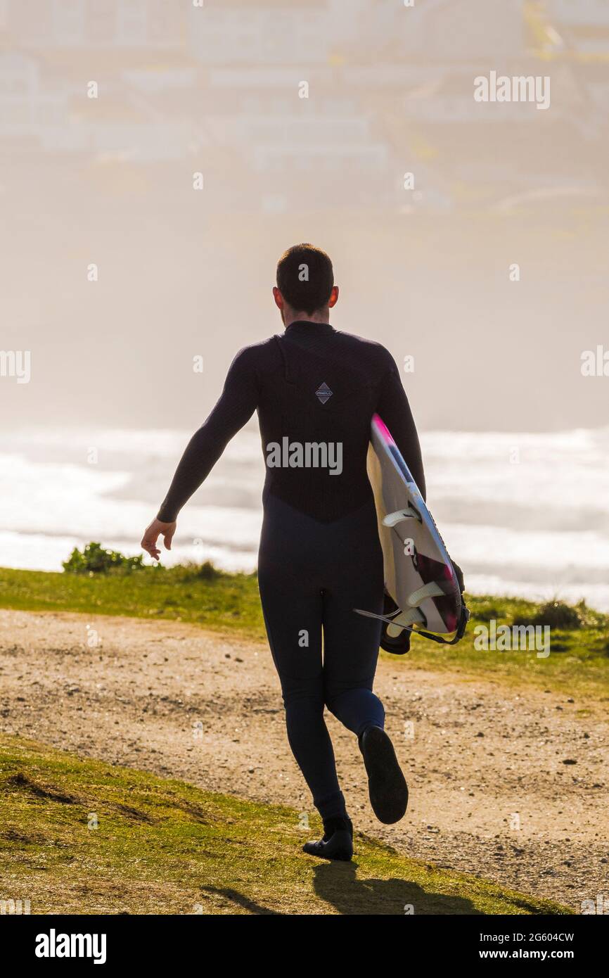The rear view of a male surfer carrying his surfboard and running along the coast path towards Fistral in Newquay in Cornwall. Stock Photo