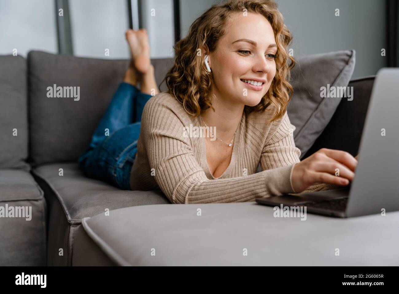 White blonde woman smiling and using laptop while lying on couch at ...