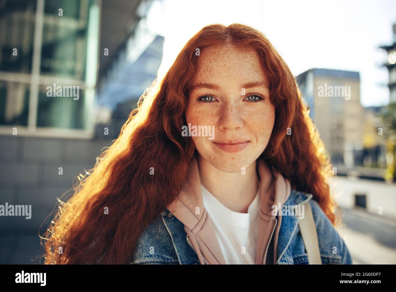 Portrait of young woman in casual wears and red dyed hair outdoors. Female student standing outside looking at camera. Stock Photo