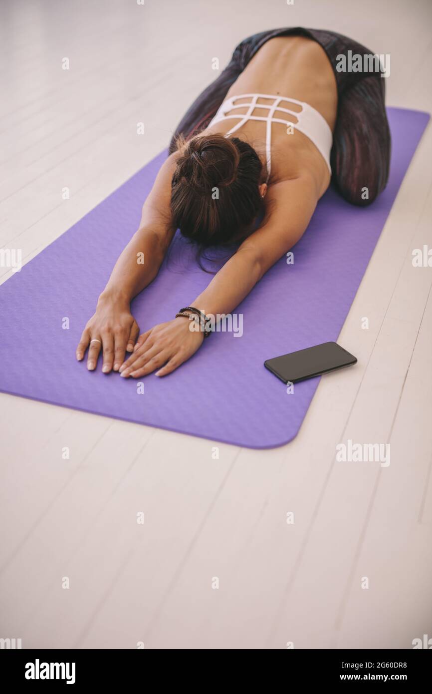 Woman doing stretching exercise on yoga mat with her cell phone on floor. fitness female performing yoga on exercise mat at gym. Child Pose, Balasana. Stock Photo