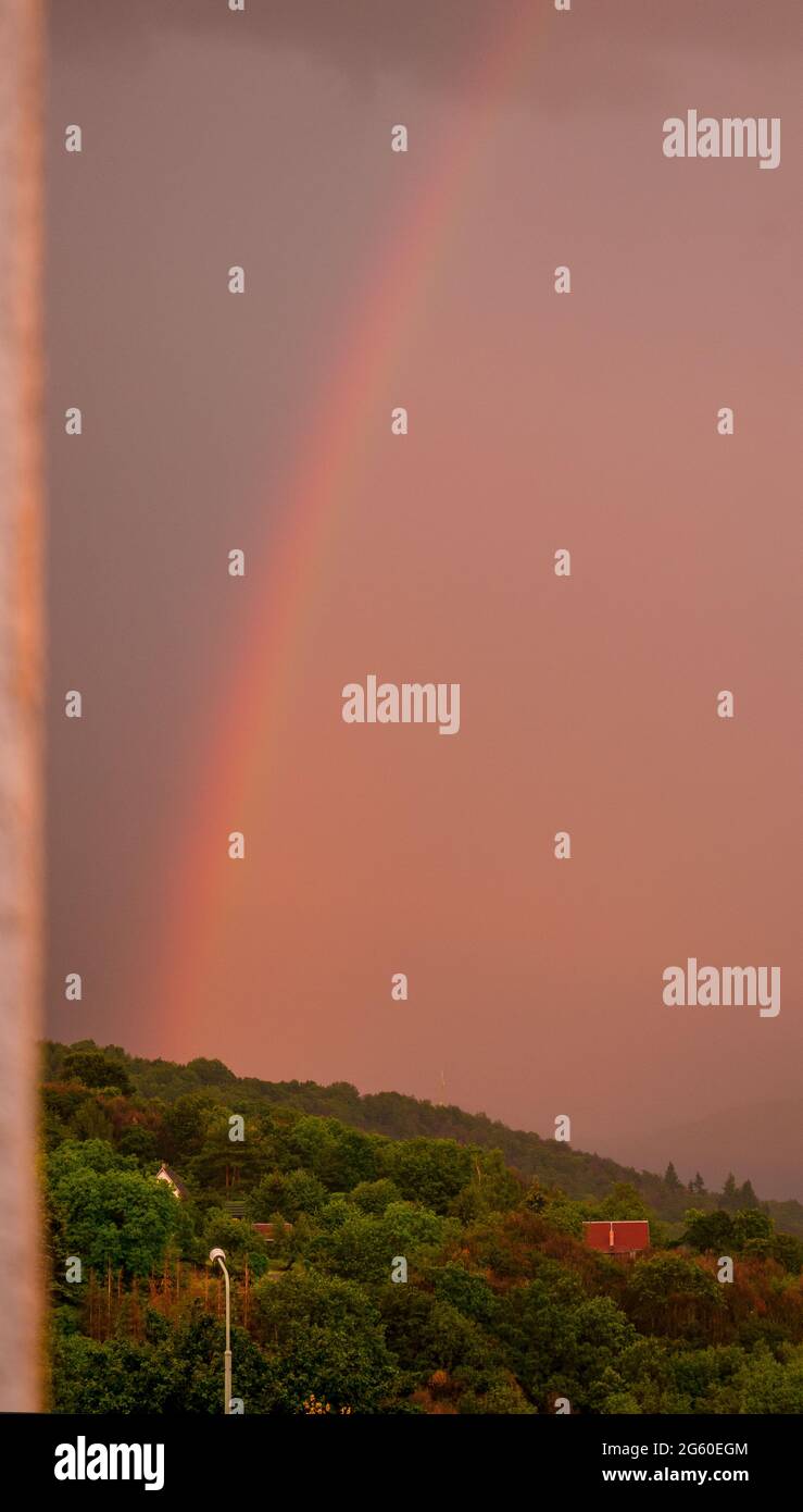Rainbow rising from the forest with dark and dense rain clouds in the background - framed by blurred wall of house Stock Photo